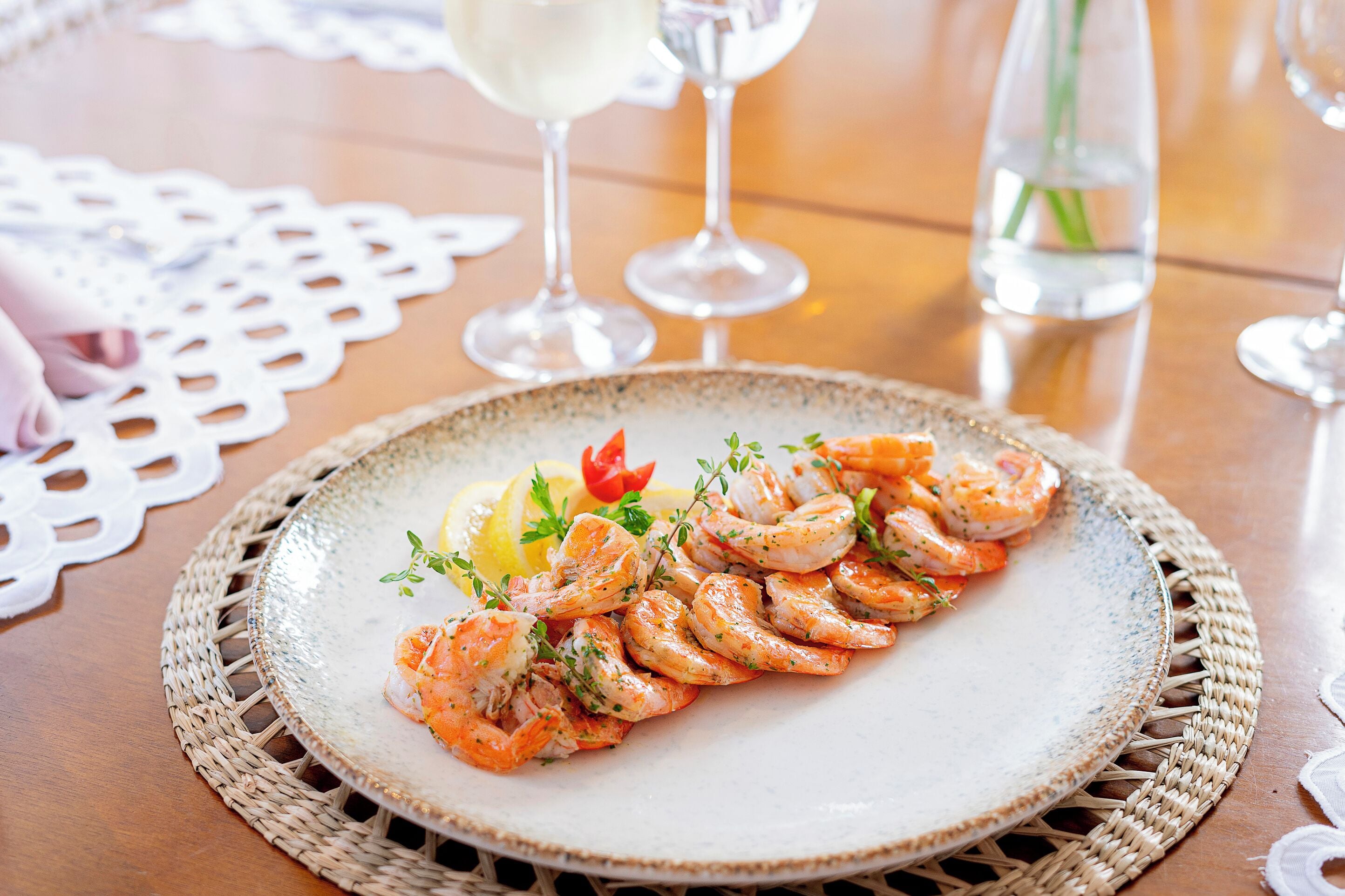 A plate of cooked shrimp garnished with herbs and lemon slices on a table with a woven placemat.