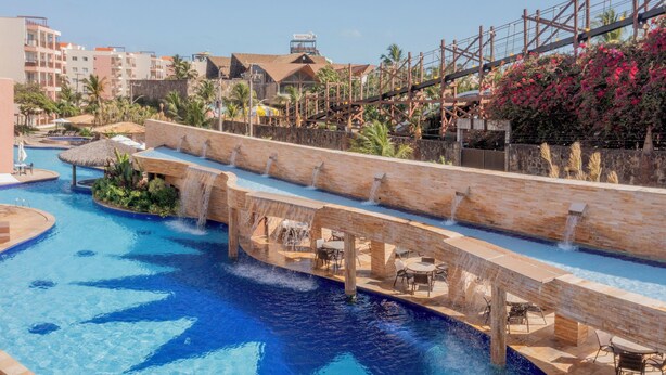 A large outdoor swimming pool with a water slide, surrounded by palm trees and a wooden bridge. - Beach Park Wellness Resort (Aquiraz)