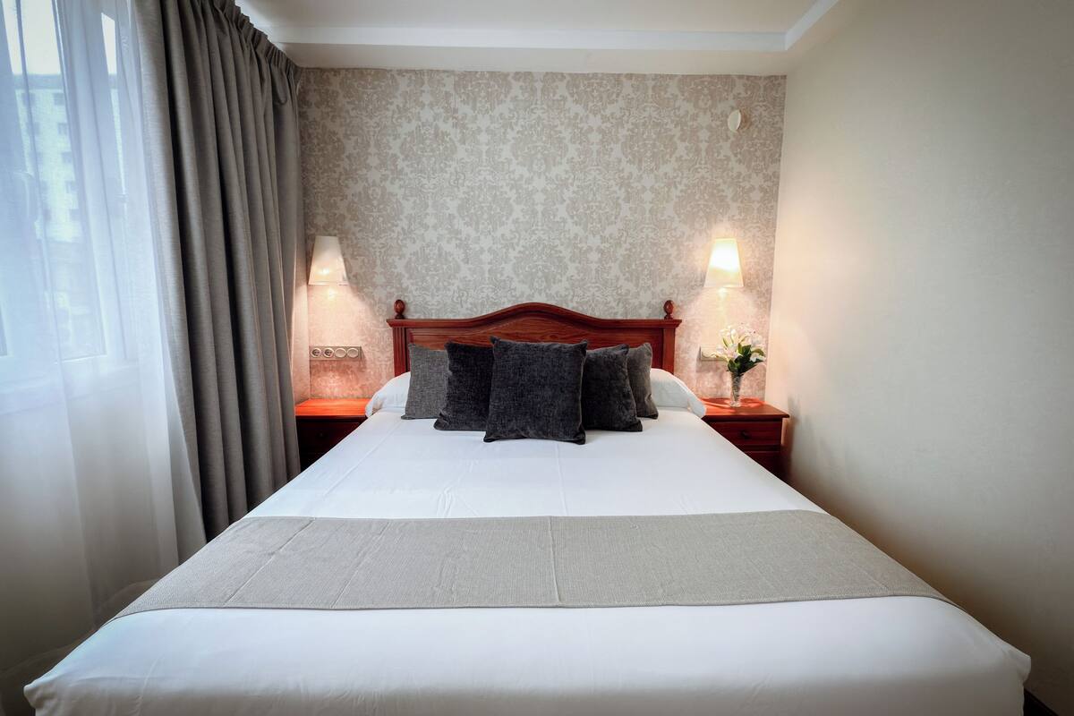 Una cama bien hecha con cabecera de madera, dos lámparas de noche y un jarrón floral sobre la mesita.