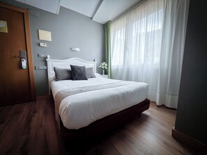 Una cama bien hecha con edredón blanco y almohadas oscuras, piso de madera, cabecera blanca, mesita de noche con un florero y ventana con cortinas translúcidas.