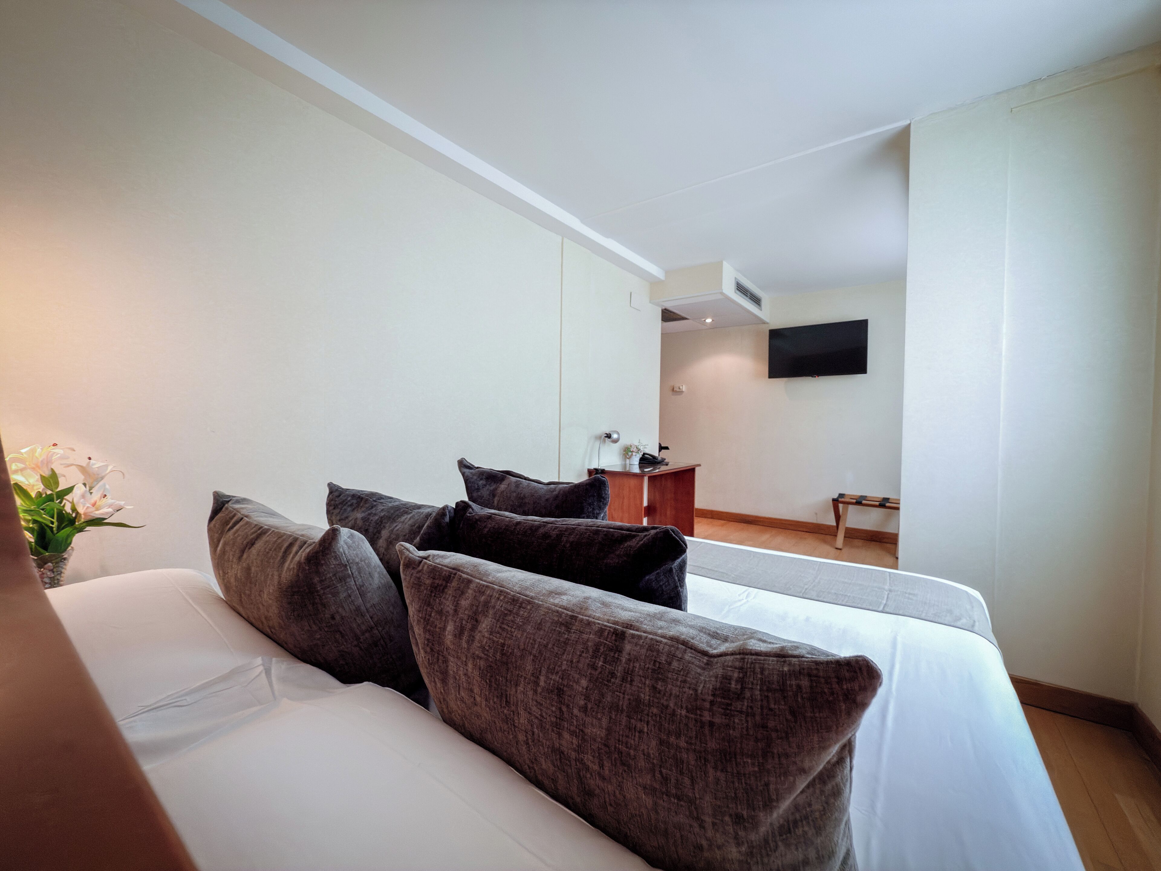 A modern hotel room with a large bed, dark brown pillows, a small desk, and a flat-screen TV mounted on the wall.