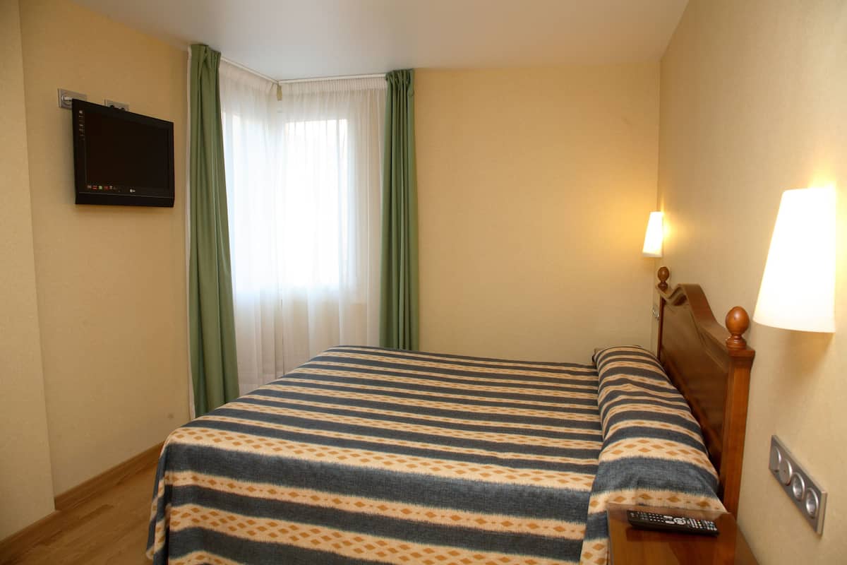 Habitación de hotel con cama, televisión montada en la pared, lámpara y control remoto sobre la mesita de noche.