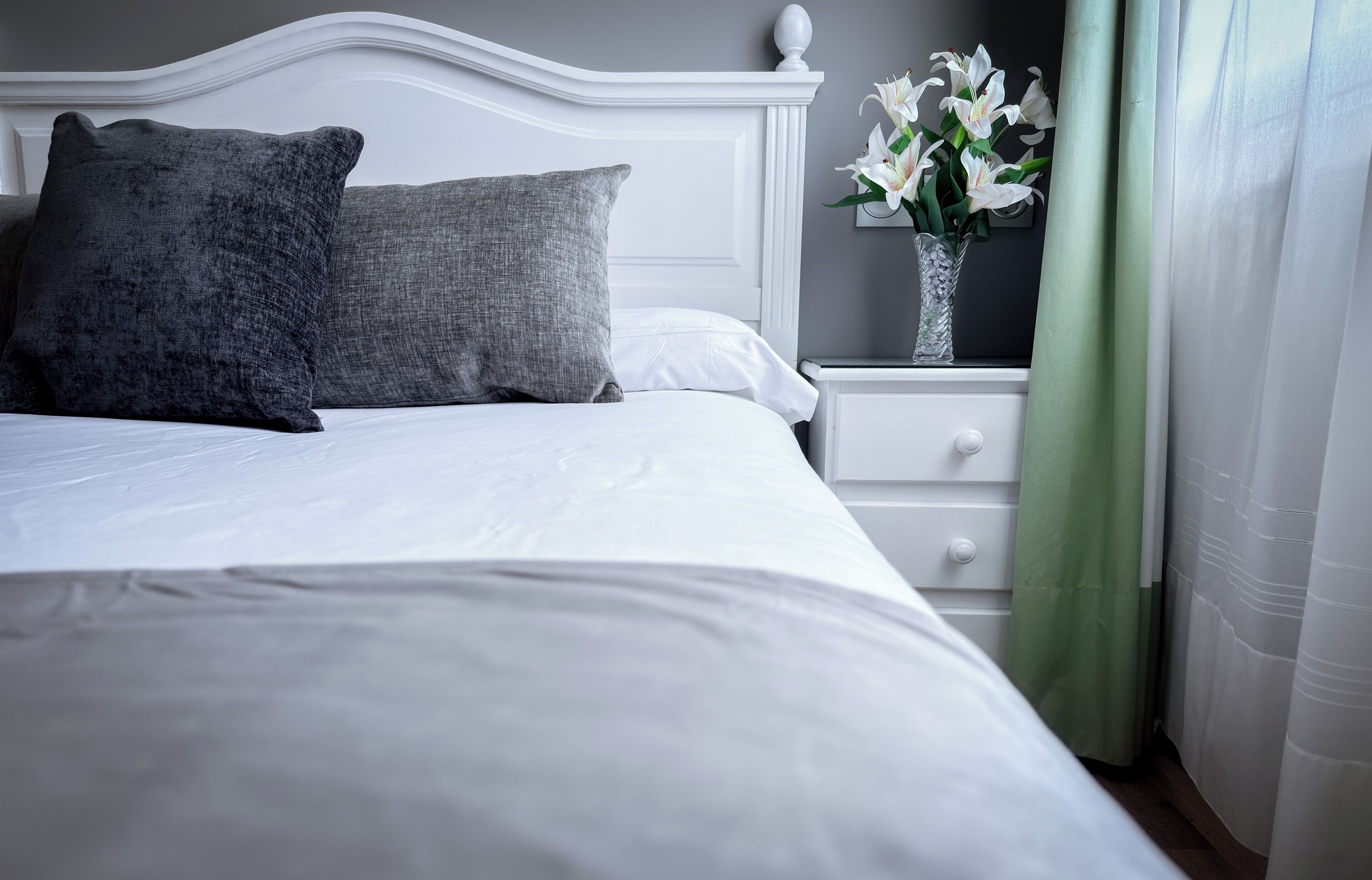 A neatly made bed with white linens and two grey pillows, a white nightstand with a vase of white flowers, and a green curtain.