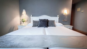Een net opgemaakt bed met witte lakens en twee kussens, een nachtkastje met een lamp, en een wandlamp.
