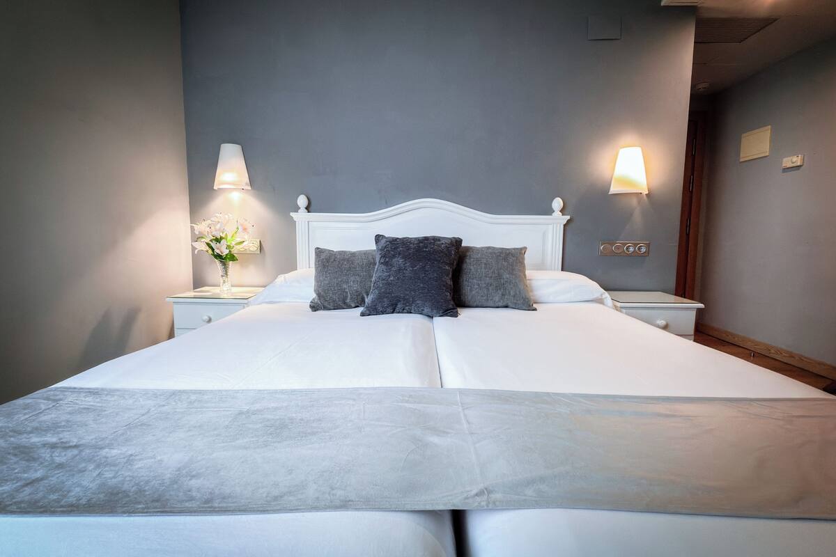 Una cama bien hecha con sábanas blancas y dos almohadas, una mesita de noche con una lámpara, y una lámpara de pared.