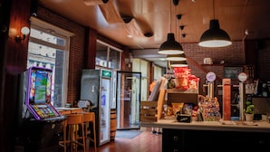 Een bar met een neonverlichte speelautomaat, een koelkast en een toonbank met snacks en drankjes.