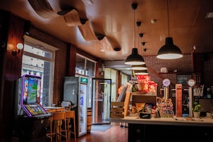 A bar area with a neon-lit arcade game, a refrigerator, and a counter with snacks and drinks.