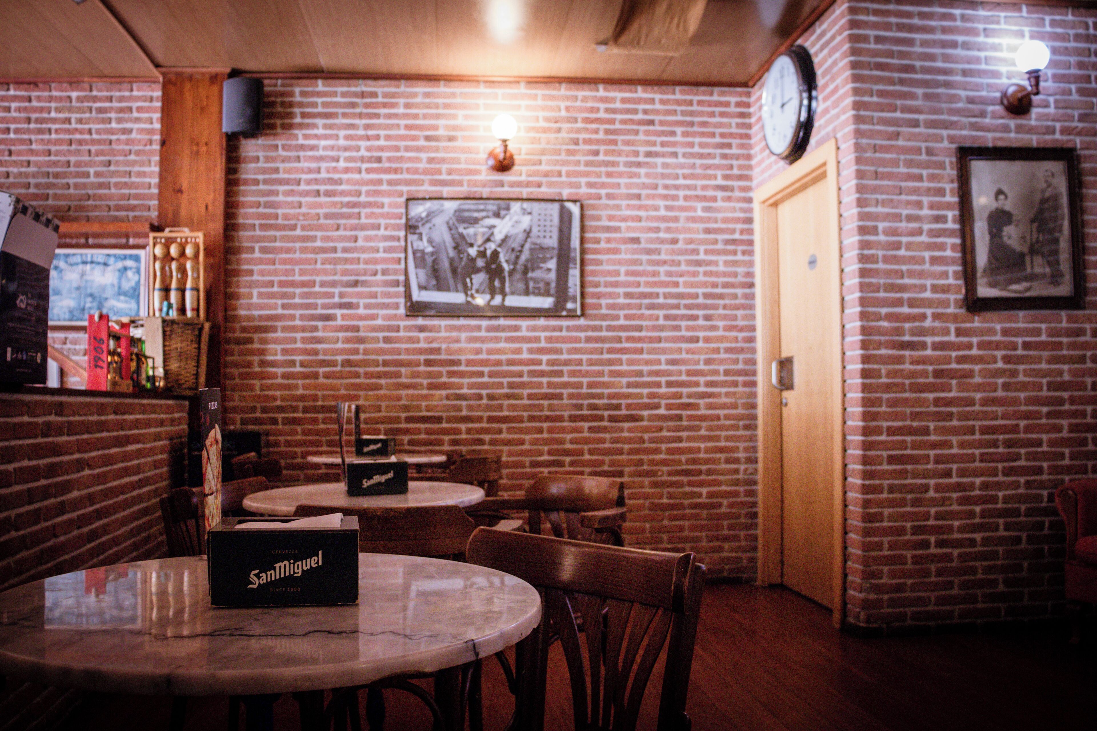 A brick-walled room with a clock, framed pictures, and a San Miguel beer box on a table.