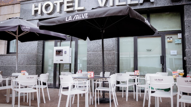 Een terras met witte stoelen en tafels, zwarte parasols en een bord met HOTEL VILLA DI LA SALVE erop.
