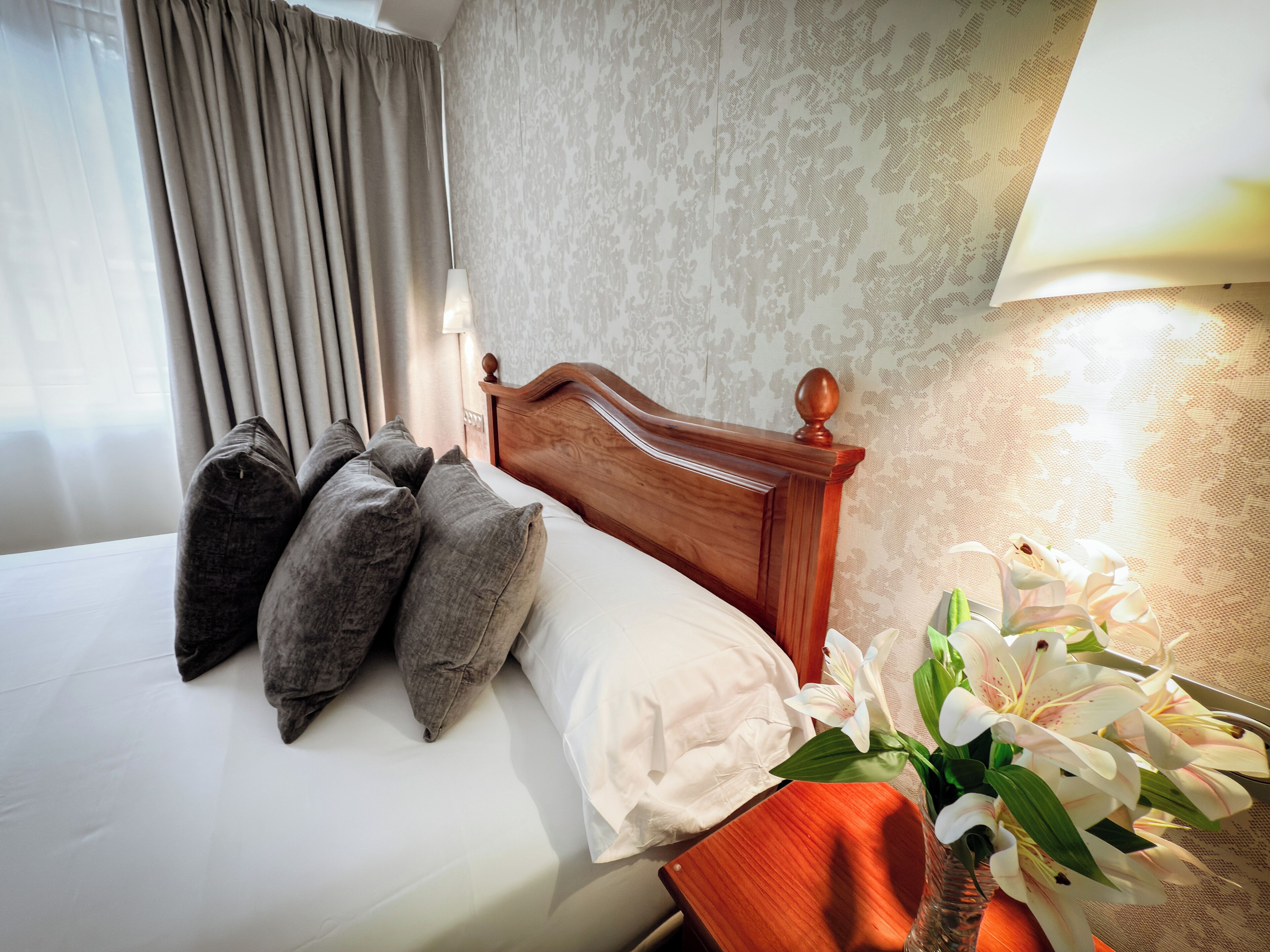 A neatly made bed with pillows, a wooden headboard, and a vase of white flowers on a nightstand.