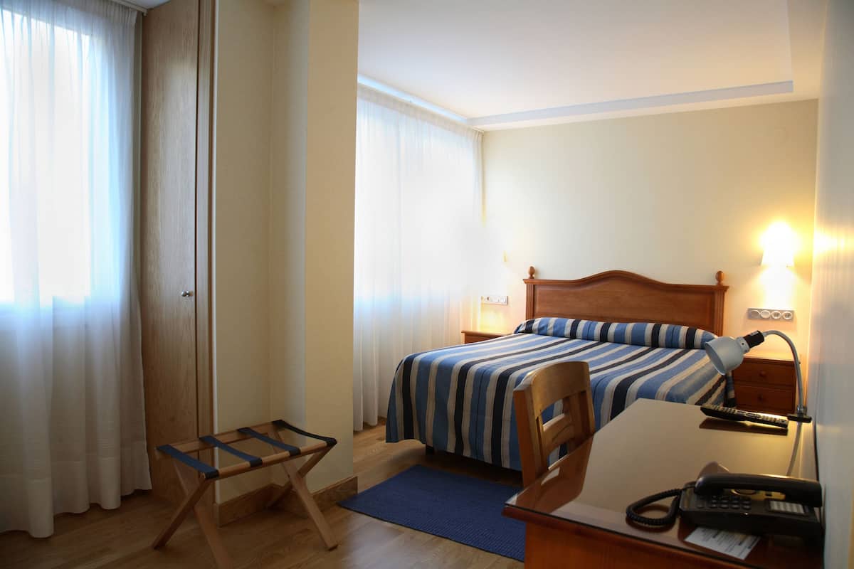Una habitación de hotel con una cama, un escritorio con teléfono, una silla, una lámpara de escritorio y una ventana con cortinas.