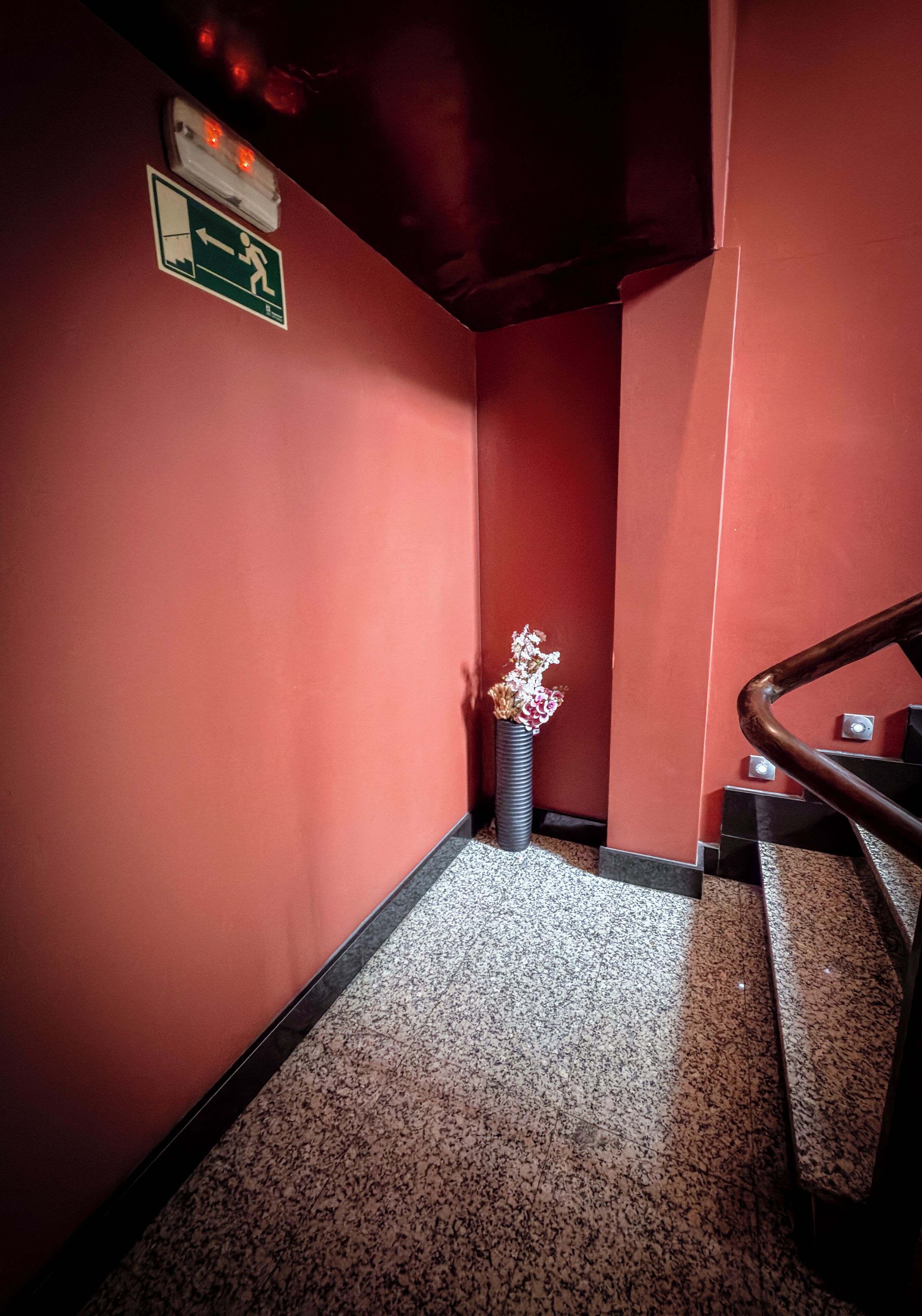 A hallway with red walls, a marble floor, a staircase with a wooden handrail, and a vase with dried flowers.