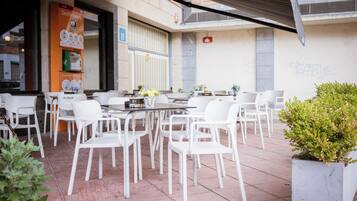 Een terras buiten met witte stoelen en tafels, een automaat en een potplant.