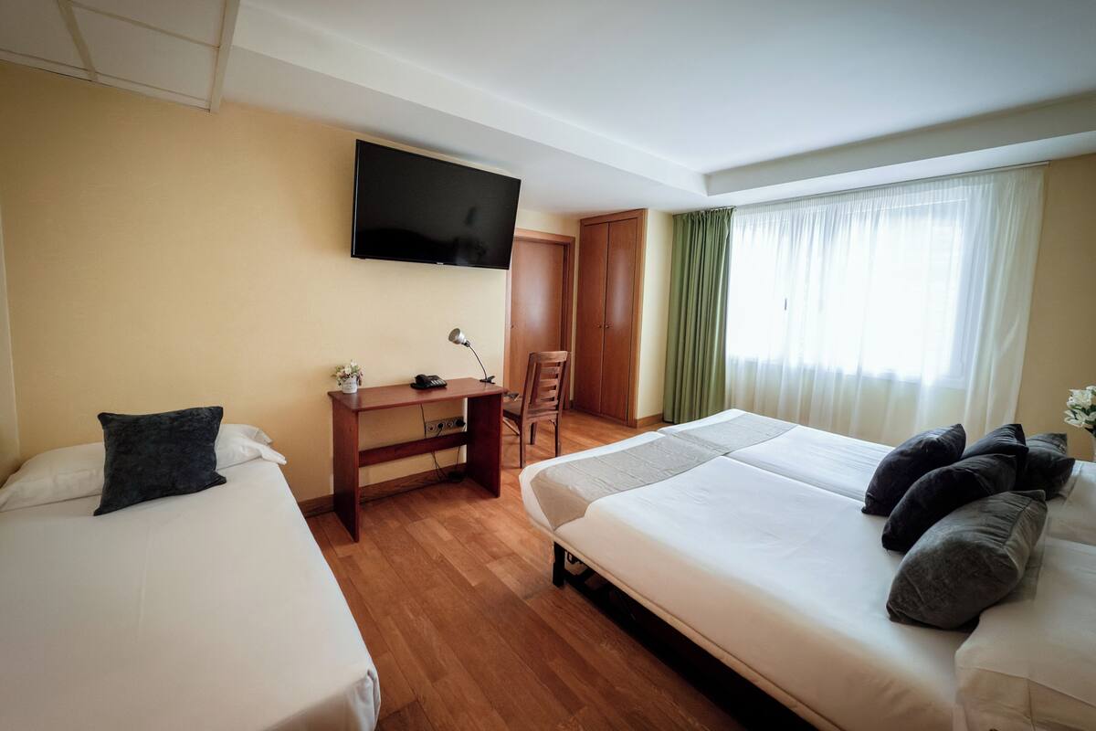 Habitación de hotel con dos camas, un escritorio con silla, televisor montado en la pared y una ventana con cortinas.