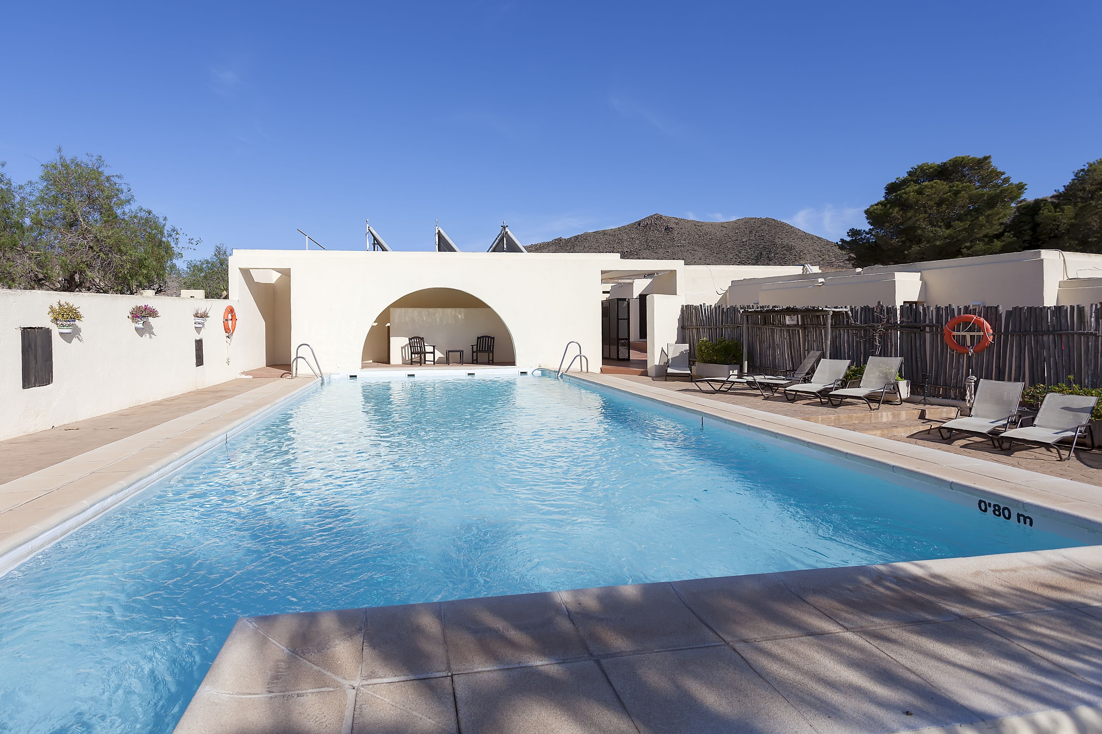 A swimming pool with a clear blue water, surrounded by lounge chairs and a wooden fence.