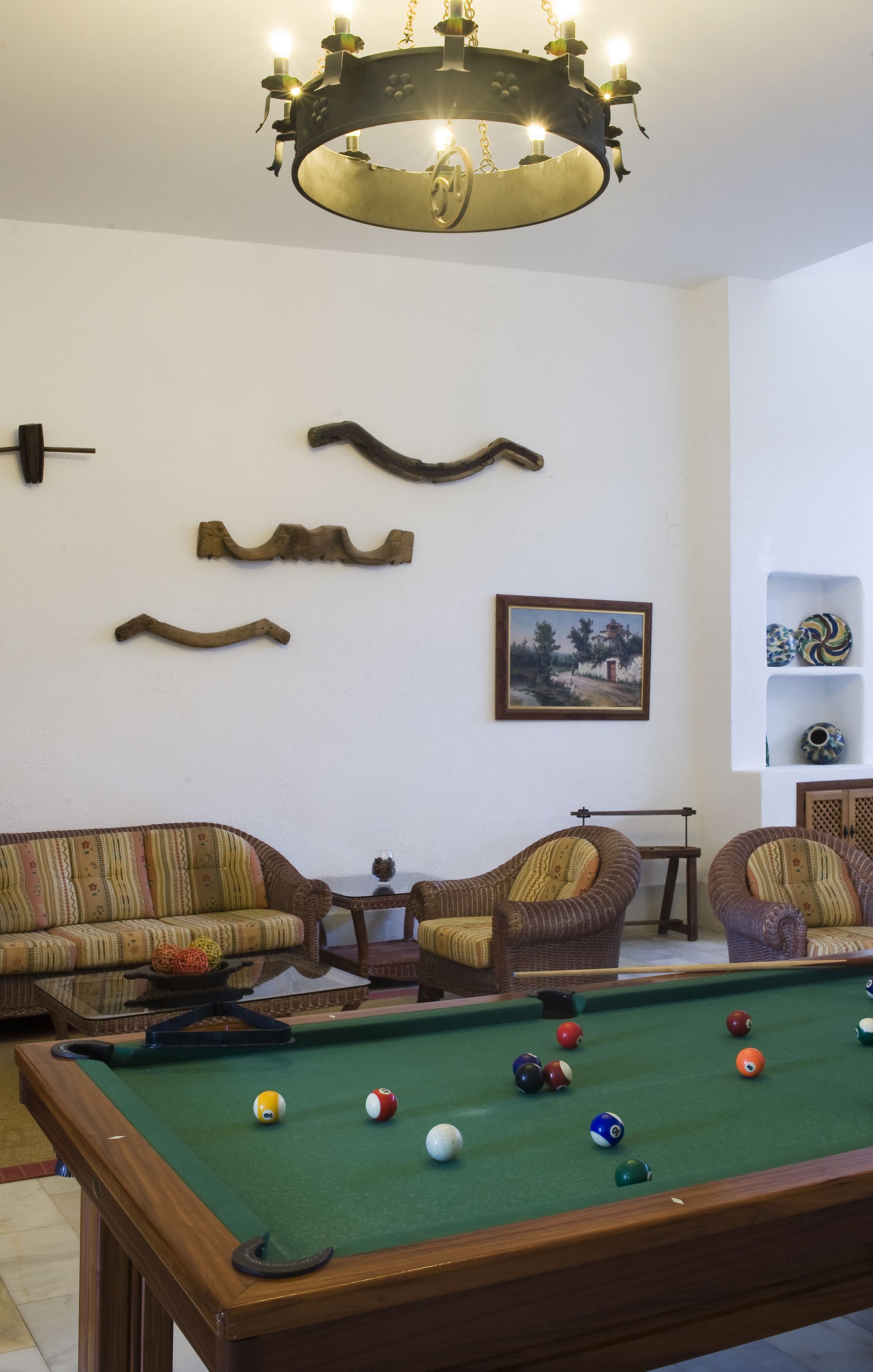 A pool table with balls, a sofa, two armchairs, a chandelier, wall decor, and a shelf with vases.