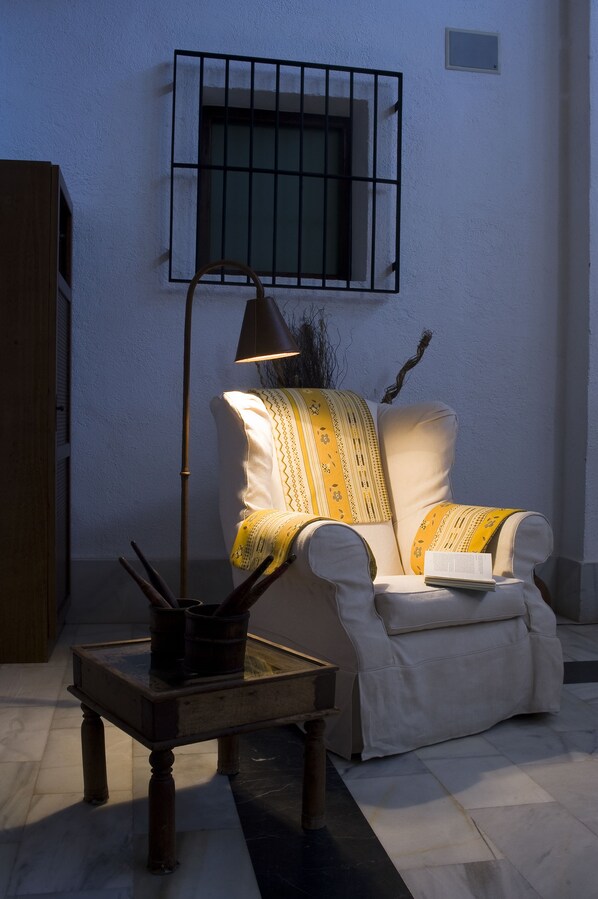Una habitación con un sillón blanco, una lámpara de pie, una mesita con un libro y una ventana con barrotes.