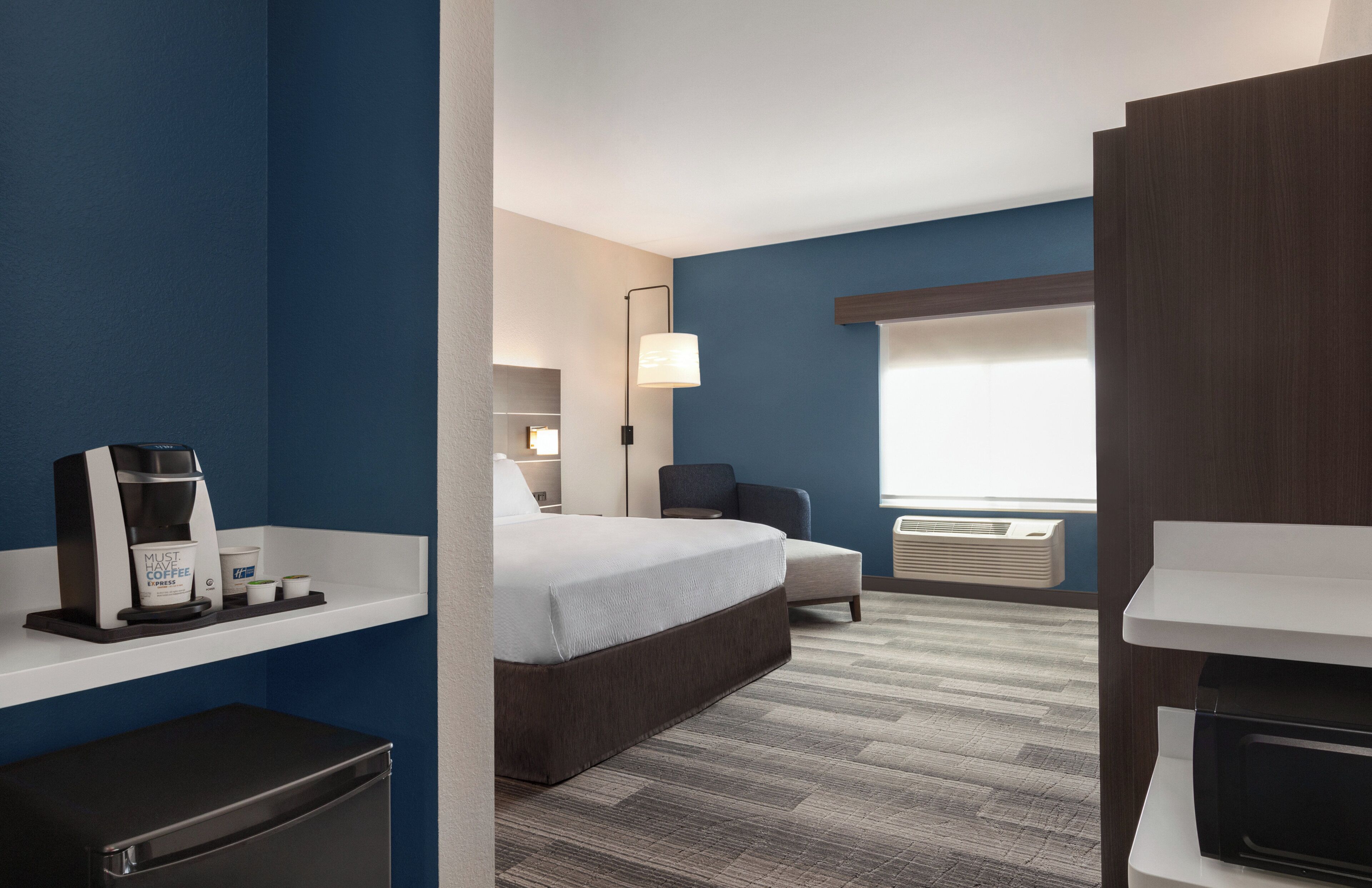 A hotel room with a bed, a chair, a desk, a coffee machine, and an air conditioning unit.