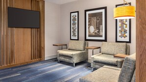 Habitación de hotel con televisor de pantalla plana montado en una pared de paneles de madera, dos sillones, un sofá y una alfombra estampada.