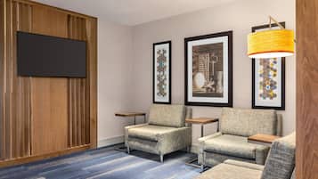 A hotel room with a flat-screen TV mounted on a wooden panel wall, two armchairs, a sofa, and a patterned carpet.