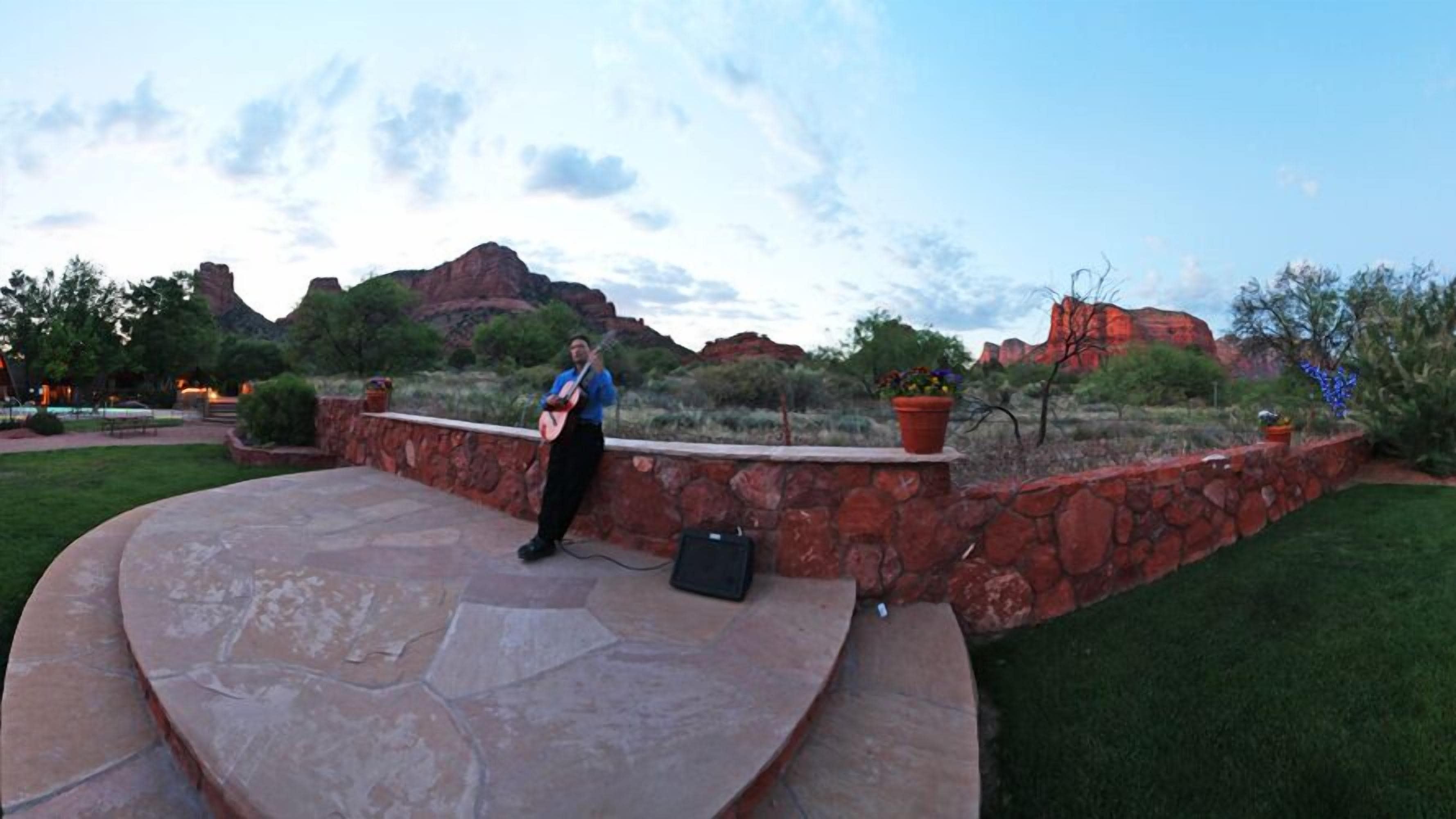 A person playing guitar on a stone stage with a stone wall backdrop and a view of red rock formations.