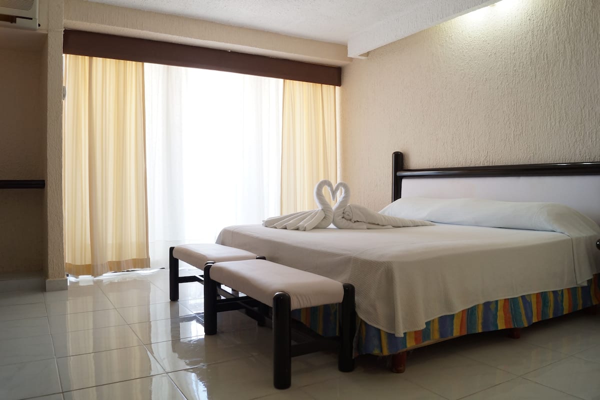 A hotel room with a bed, bench, curtains, and a wall-mounted air conditioner.