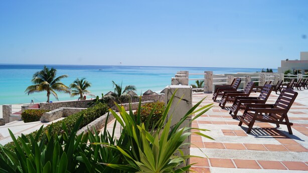 Una terraza con barandillas de piedra, palmeras y sillones de descanso con vista a una playa.