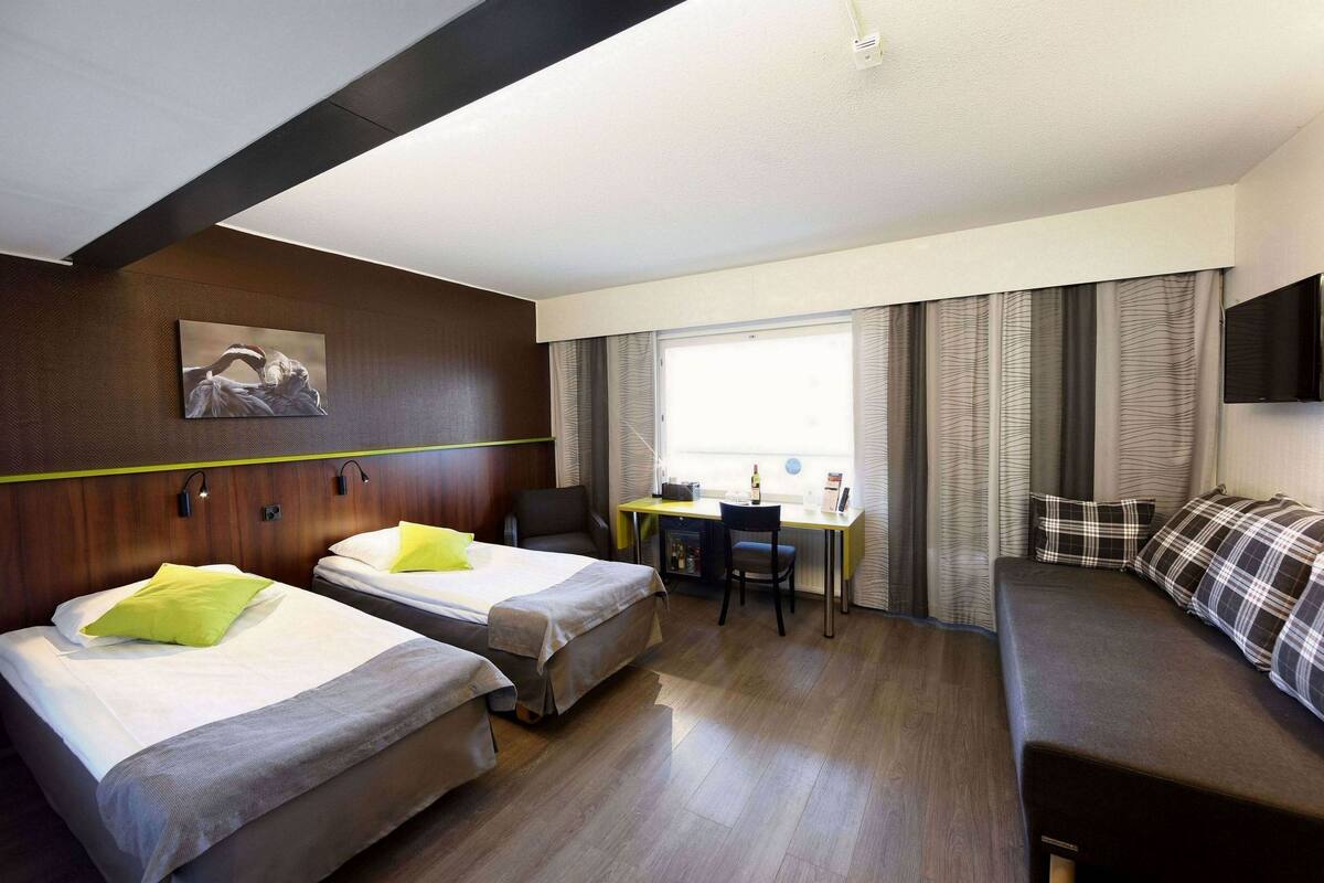 A hotel room with two beds, a desk, a chair, a sofa, a painting on the wall, and a large window with curtains.