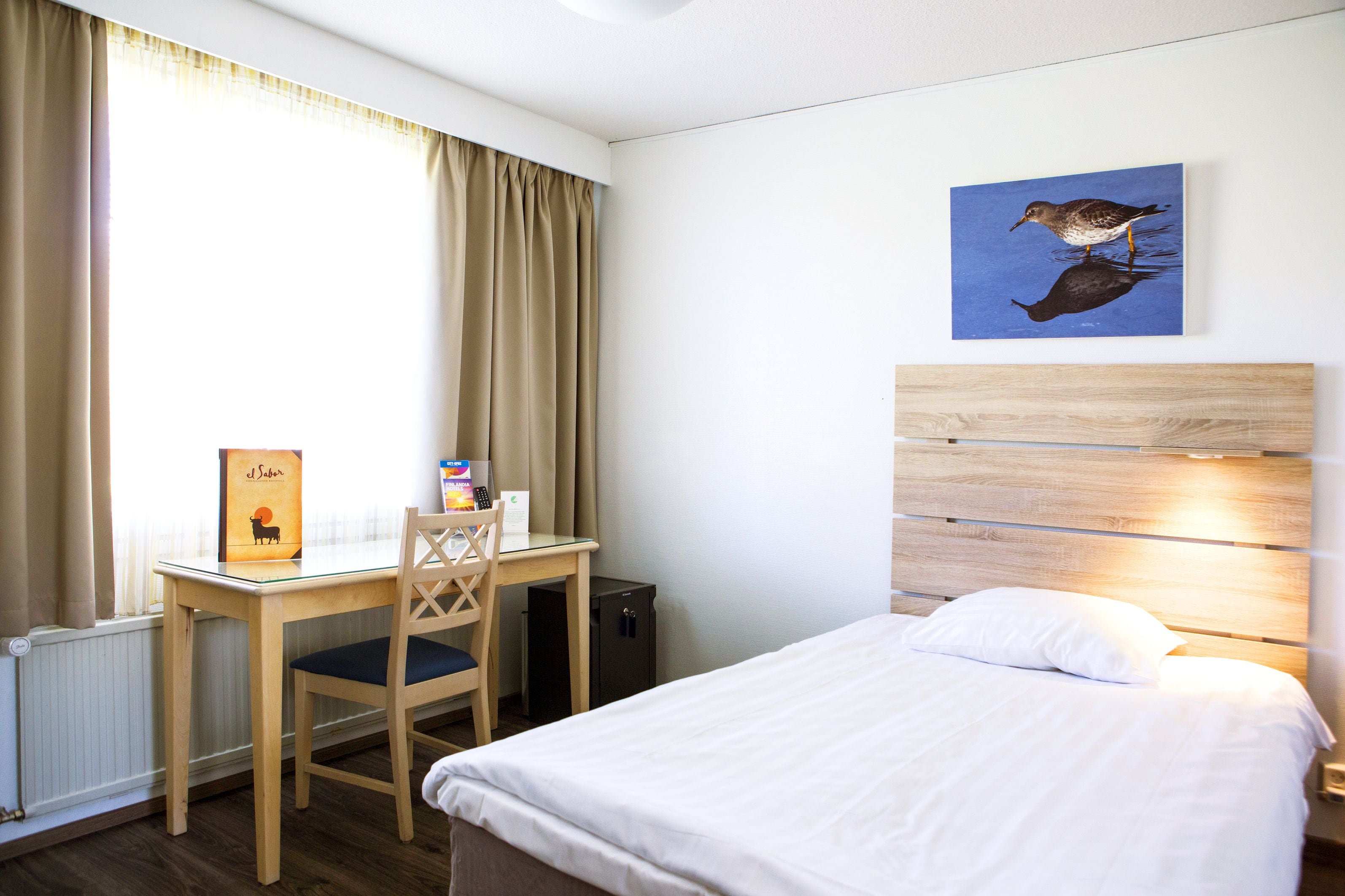 A hotel room with a bed, a desk with a chair, a radiator, a picture on the wall, and a window with curtains.
