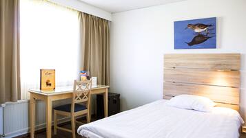 A hotel room with a bed, a desk with a chair, a radiator, a picture on the wall, and a window with curtains.