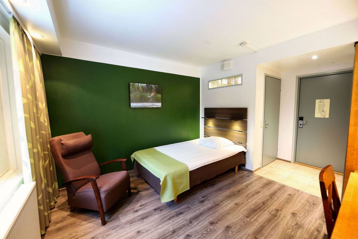 A bedroom with a bed, a chair, a green wall, and a wooden floor.