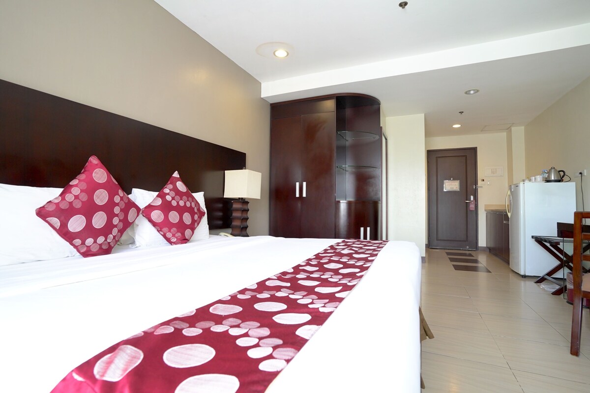 A hotel room with a large bed, a wooden wardrobe, a desk, and a kitchenette.
