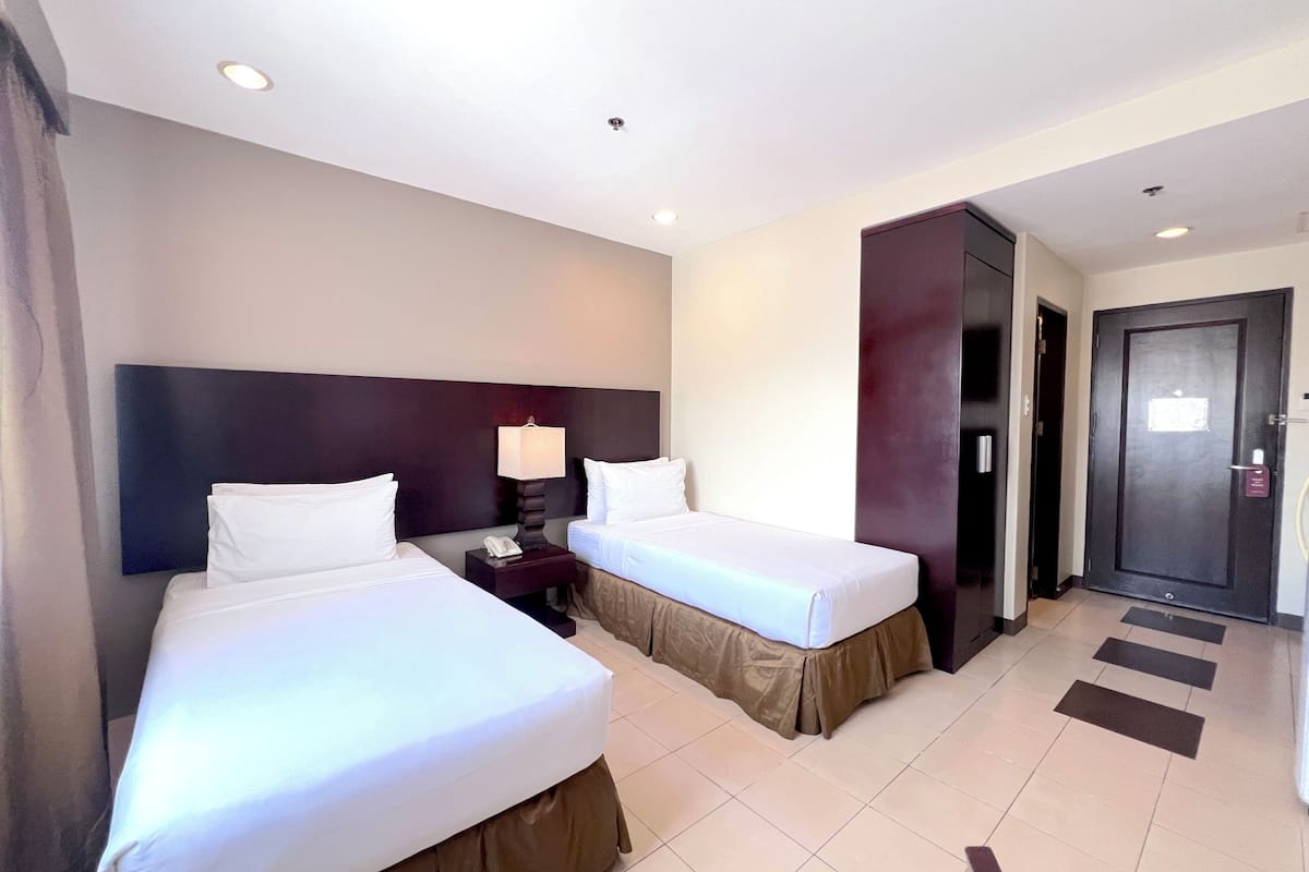 Habitación de hotel con dos camas, una mesita de noche con lámpara, una puerta y suelo de baldosas.