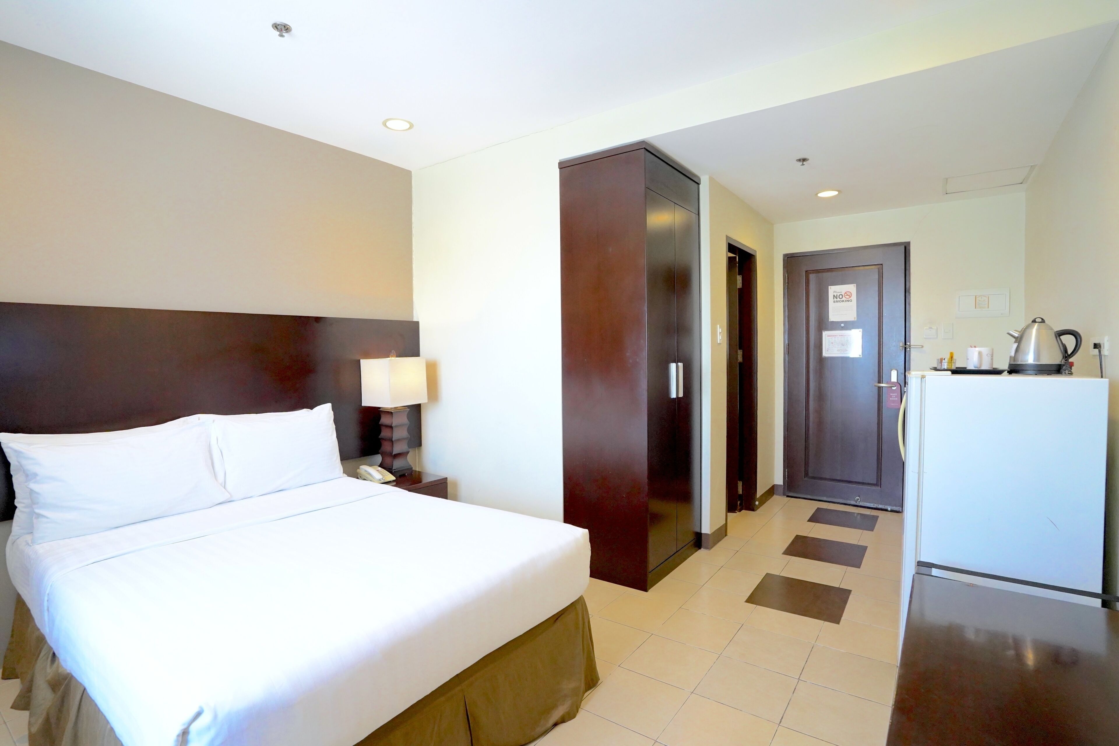 Una camera d'albergo con un letto, un comodino, una lampada, un armadio, una porta e un frigorifero.
