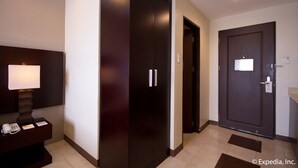 A hotel room with a dark wooden wardrobe, a desk with a telephone and a lamp, and a door with a sign. - Alpa City Suites (Mandaue)