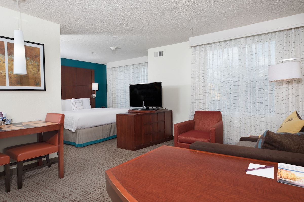 Quarto de hotel com cama, televisão, secretária, cadeira, sofá e uma janela grande com cortinas.