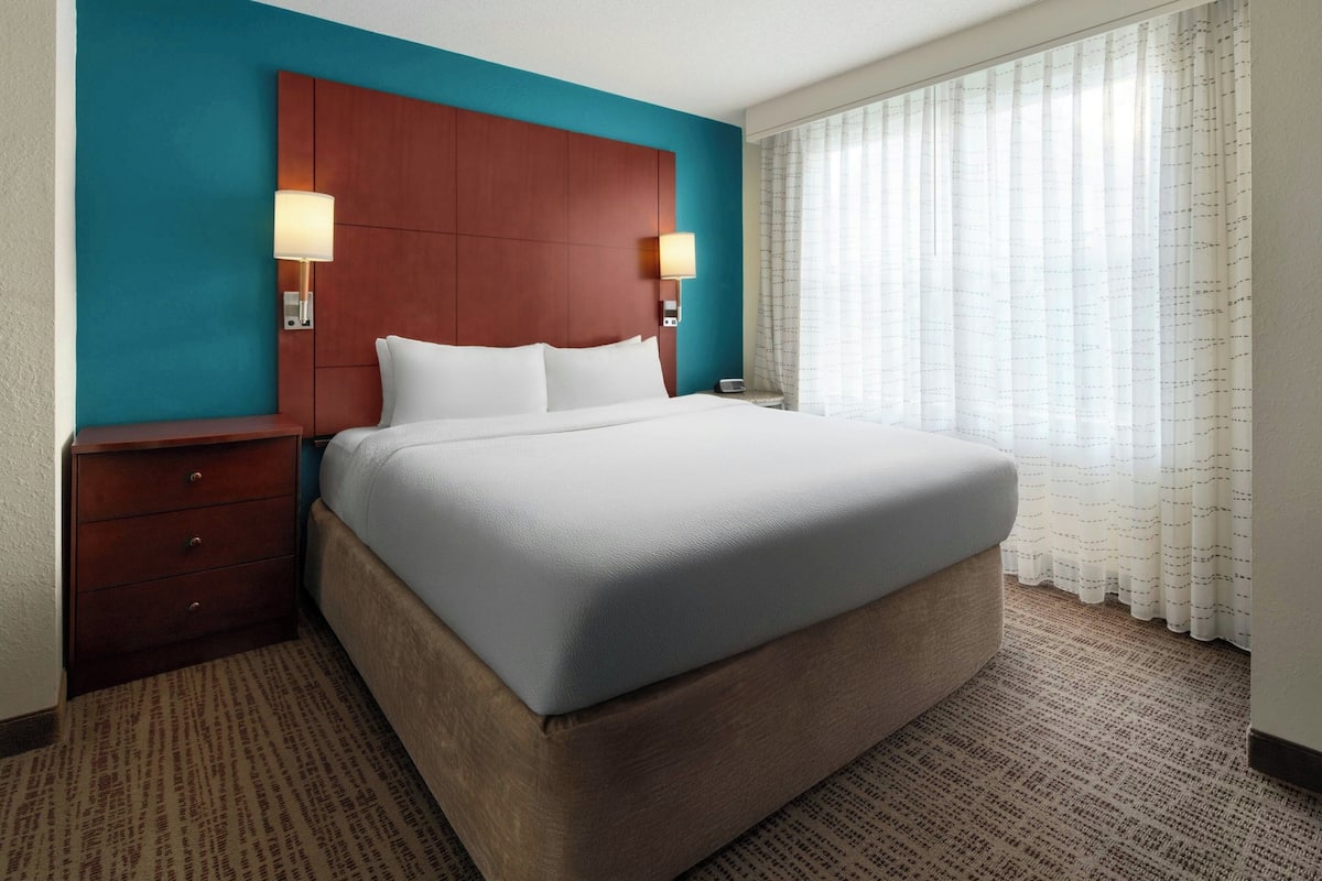 Quarto de hotel com uma cama grande, cabeceira de madeira, duas mesas de cabeceira com abajures e uma janela com cortinas leves.