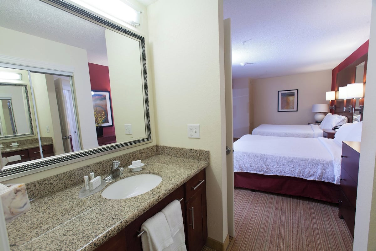 Quarto de hotel com um espelho grande no banheiro, pia e cama com lençóis brancos.