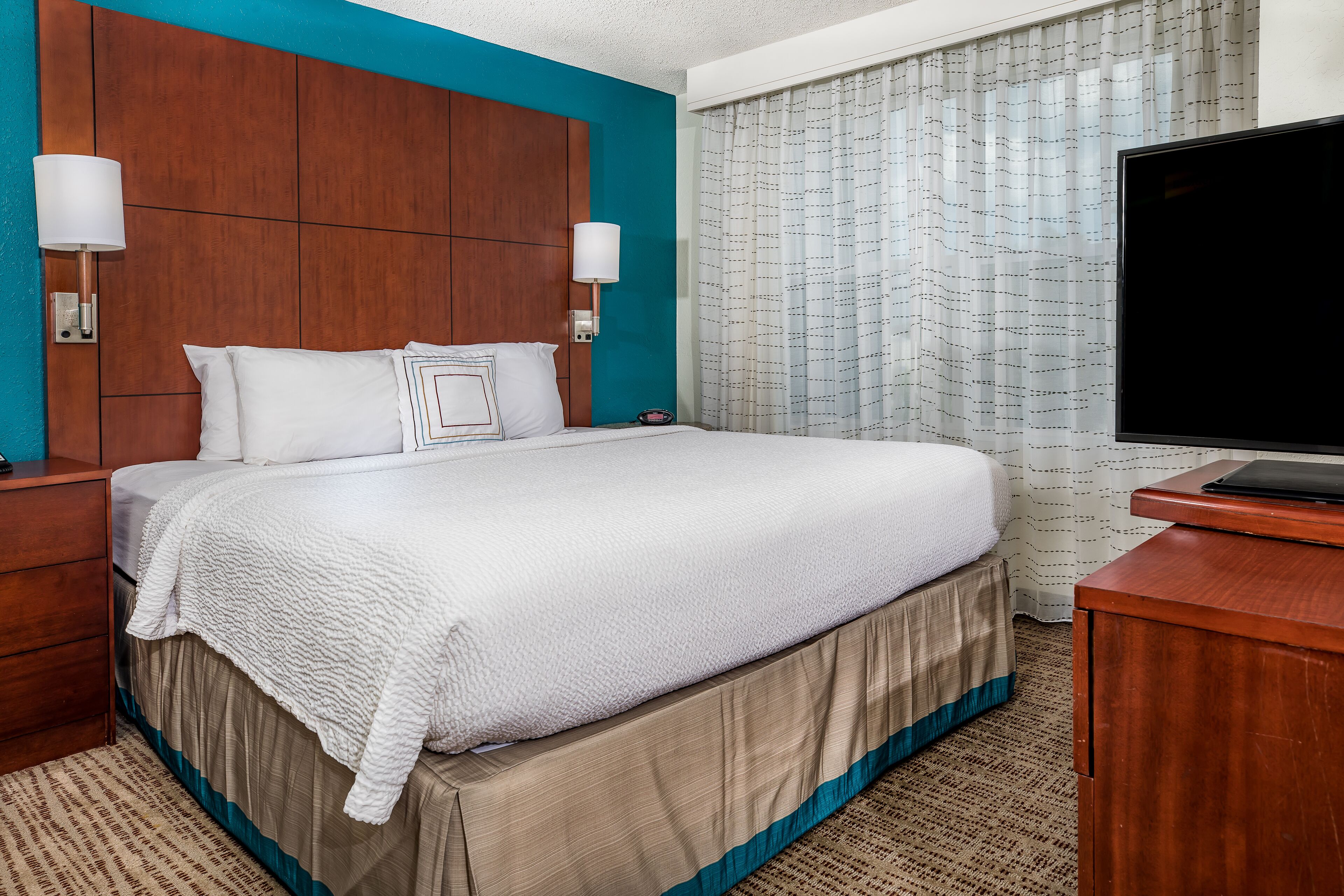 A hotel room with a large bed, wooden headboard, two bedside lamps, a flat-screen TV, and a window with patterned curtains.