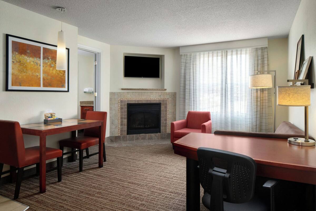 Quarto de hotel com lareira, uma TV de tela plana acima dela, área de jantar com mesa e cadeiras, e um sofá.