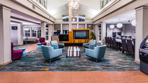 Un vestíbulo amplio con techo alto, zona de estar central con sillones y una mesa de centro, televisor de pantalla plana y comedor con mesas y sillas.