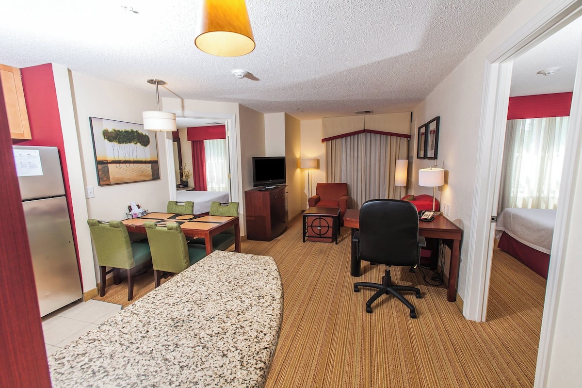 Quarto de hotel com cama, escrivaninha, cadeira, televisão e área de refeições com cadeiras e mesa.