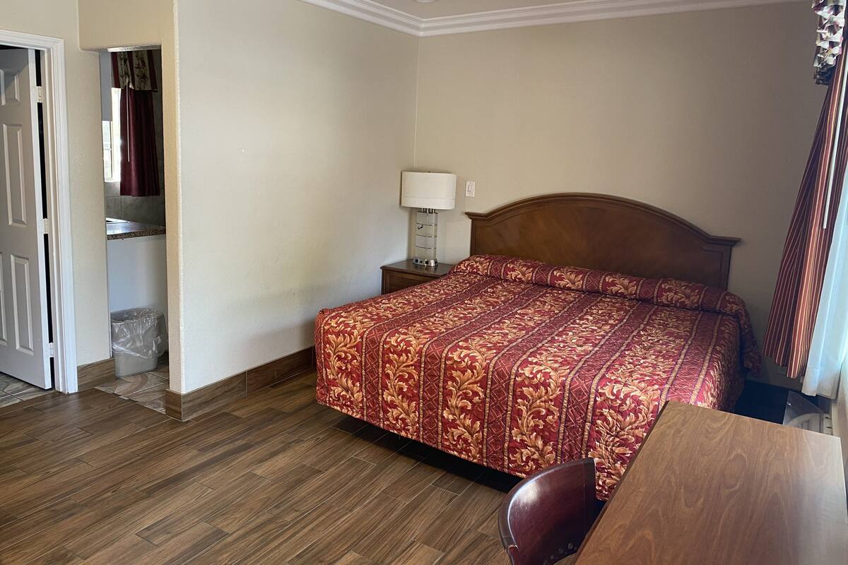 Una habitación de hotel con suelo de madera, una cama con cubrecama estampado, una mesita de noche con lámpara y un baño visible a través de una puerta abierta.