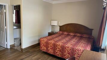 A hotel room with a wooden floor, a bed with a patterned bedspread, a nightstand with a lamp, and a bathroom visible through an open door.