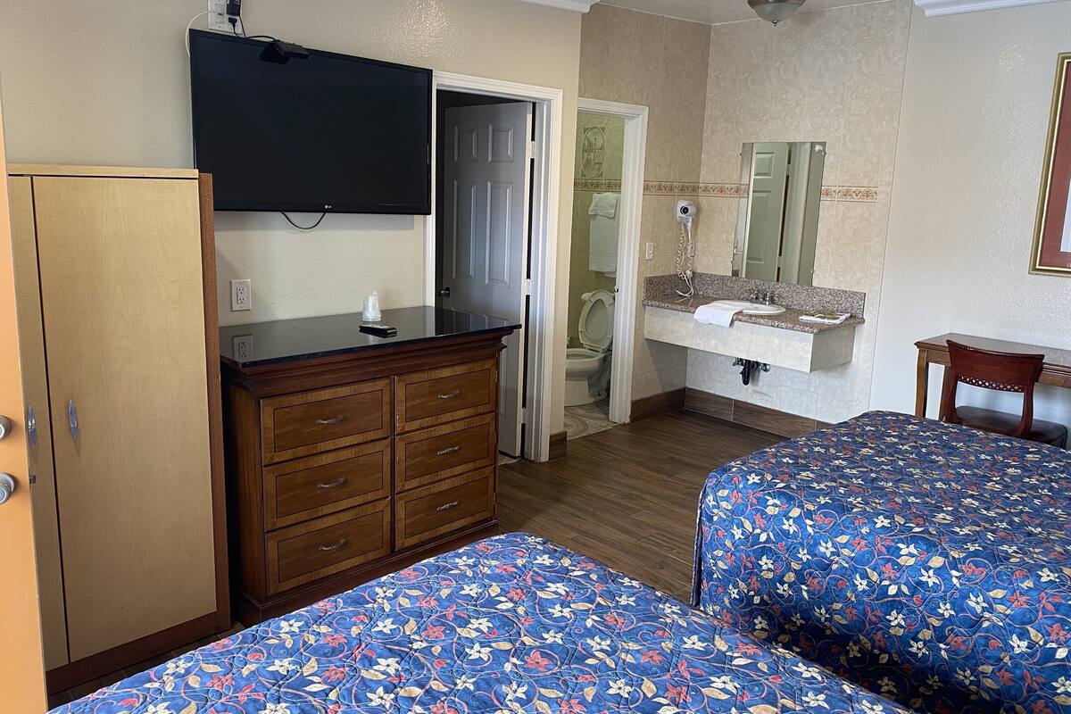 Habitación de hotel con dos camas, un aparador de madera, un televisor, un baño con lavamanos y espejo, y una mesita.