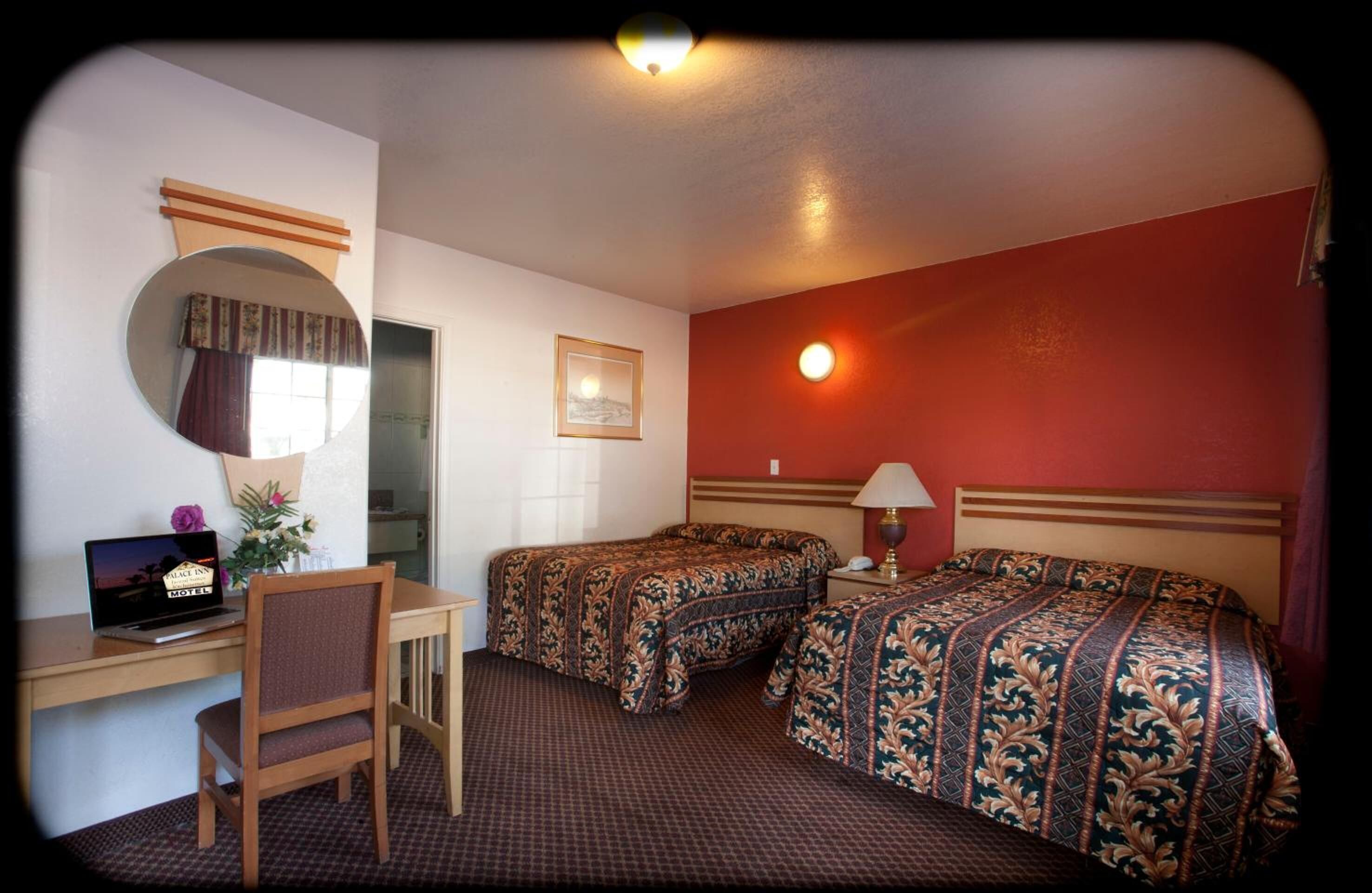 A hotel room with two beds, a desk with a laptop, a chair, a mirror, a lamp, and a framed picture on the wall.