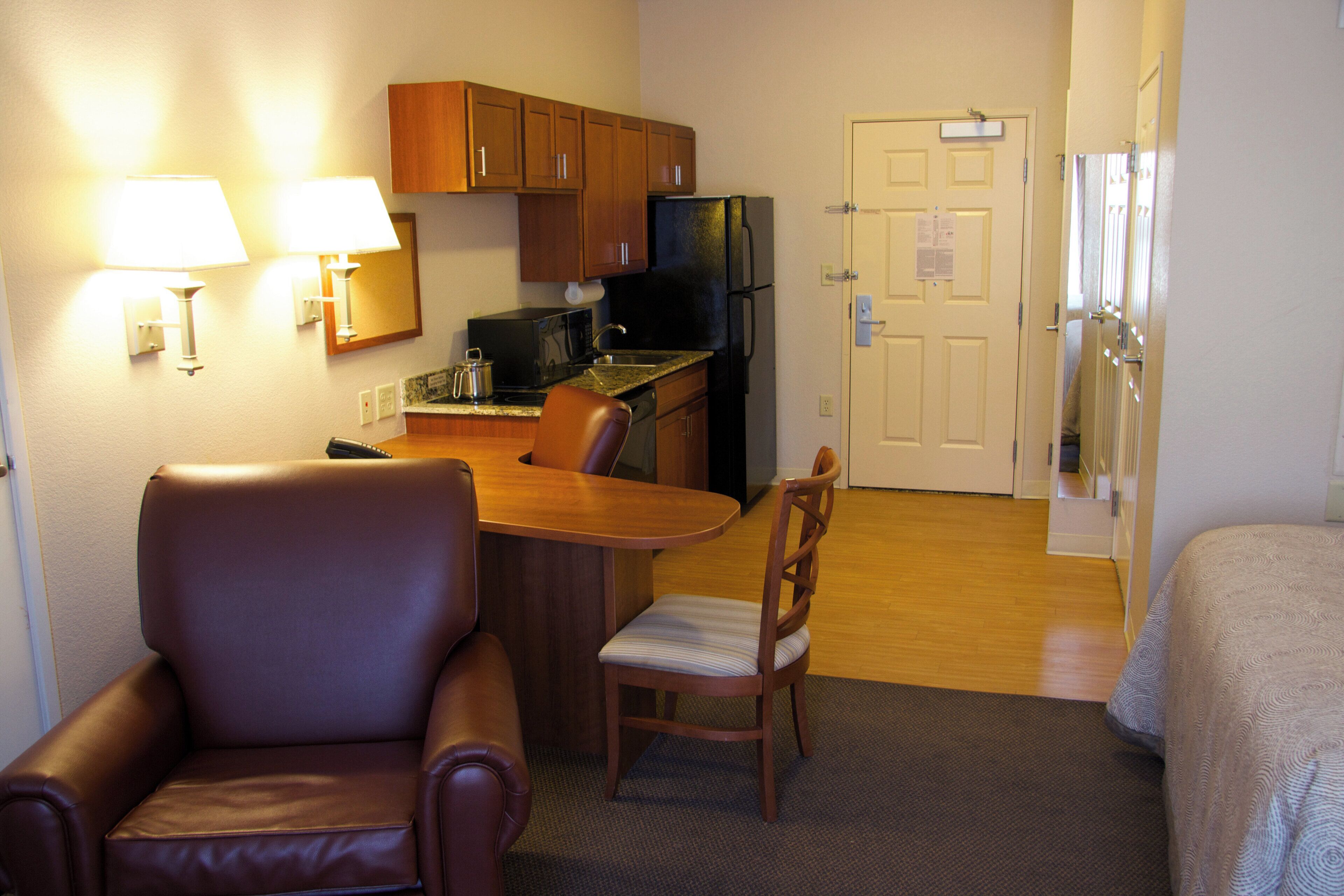 A compact hotel room with a kitchenette, a dining area with a table and chairs, a bed, and a door.
