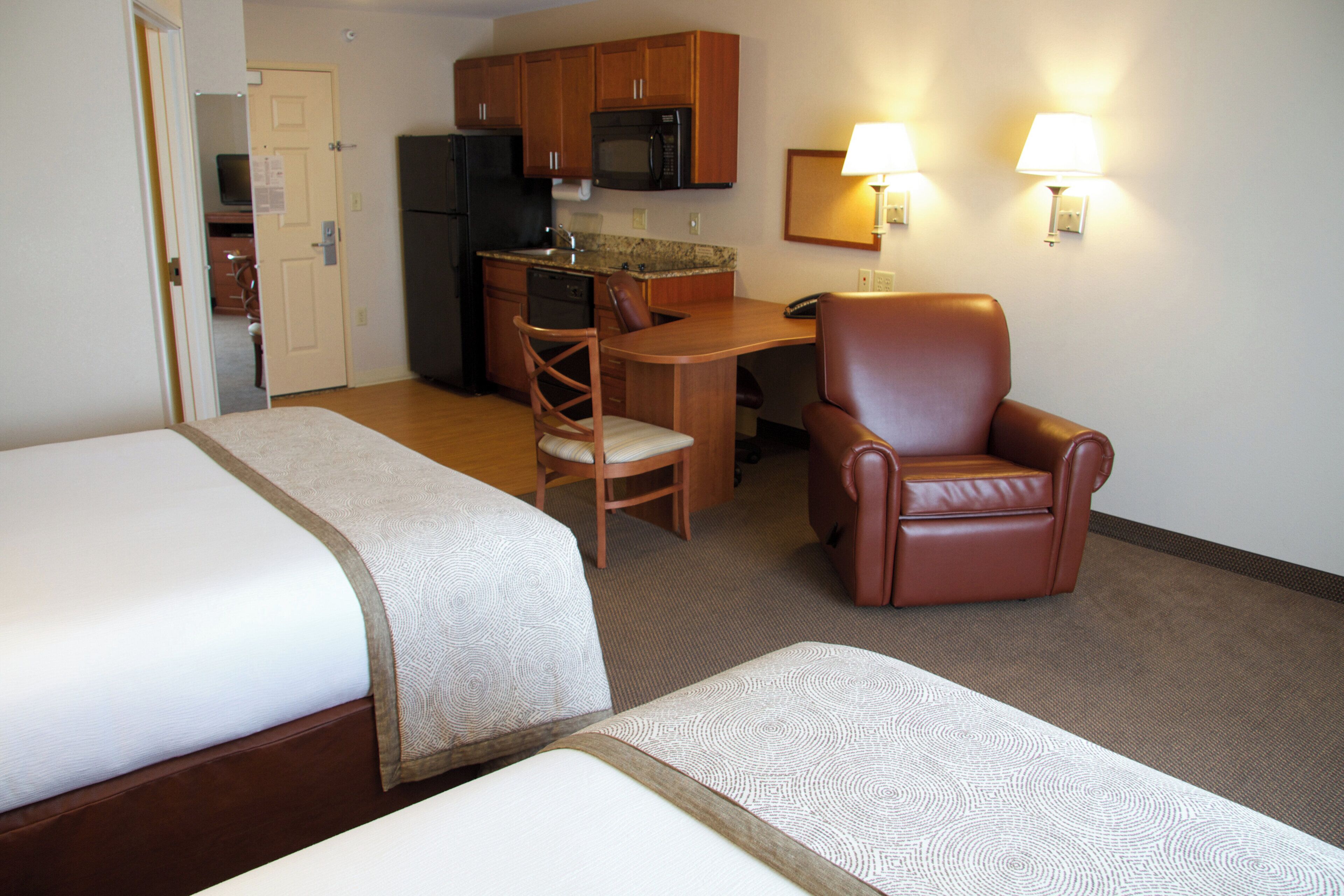 A hotel room with two beds, a kitchenette, a dining area, and a seating area with a chair and a lamp.