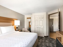 Habitación de hotel con cama, mesita de noche, lámpara, silla, armario y puerta que da a otra habitación.