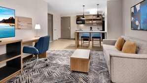 A modern hotel room with a flat-screen TV, a desk with a chair, a sofa, a coffee table, a patterned rug, and a kitchenette in the background.